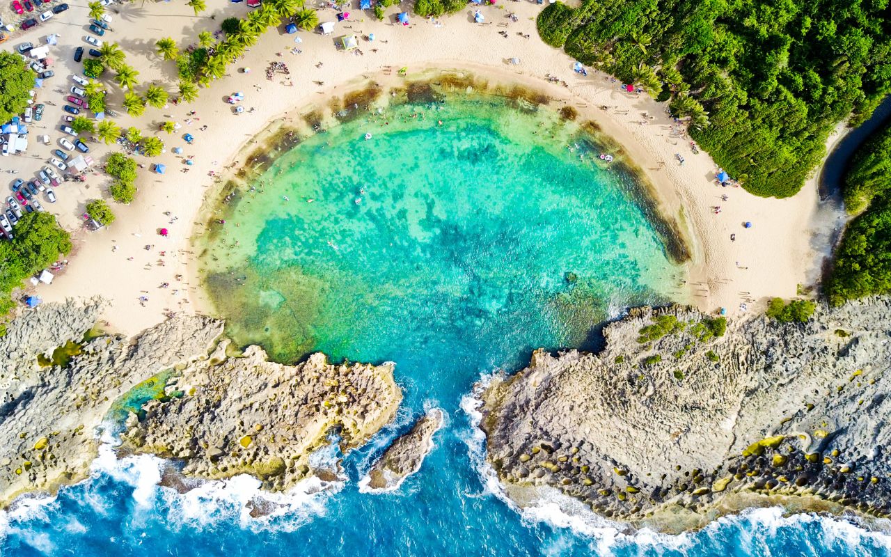 Birds-eye-view of turquoise water in a circular tidal pool with a sandy beach and rocky shoreline and people lounging on the beach