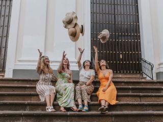 A group of four women throw their hats in the air while sitting on the steps in front of a building 